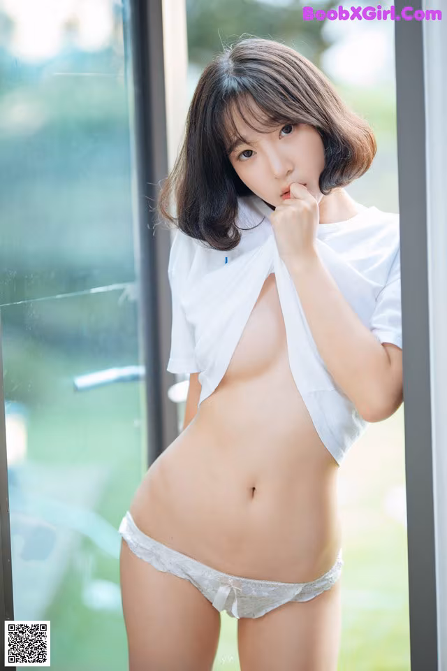 A woman in a white shirt and panties posing by a window.